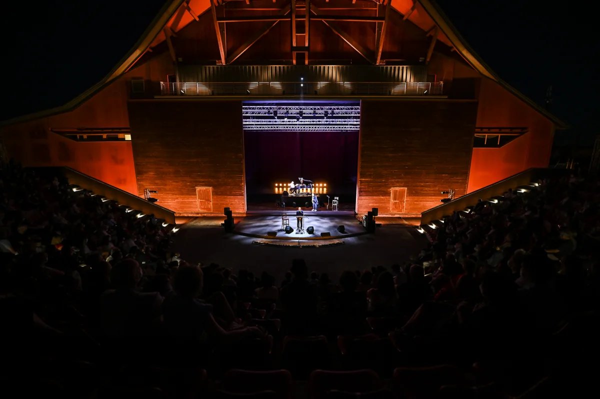 L'immagine dell'Anfiteatro Julian Beck del Teatro Era di Pontedera: presenta una vista accattivante di un palcoscenico teatrale aperto, splendidamente illuminato e allestito per uno spettacolo che può essere organizzato anche da aziende private. Il palcoscenico è adornato con colori ricchi e vibranti e luci intricate, che creano un'atmosfera invitante per il pubblico. L'illuminazione proietta una luce calda, esaltando i dettagli della scenografia e attirando l'attenzione sui punti focali del palco. La composizione complessiva trasmette un senso di anticipazione ed eccitazione, riflettendo il ruolo del teatro aperto come spazio di creatività ed espressione per poter organizzare eventi aziendali.