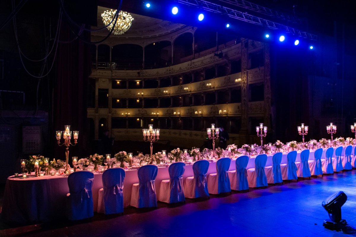 Una scena vivace e invitante, probabilmente pensata per promuovere l'organizzazione di eventi. Mostra una tavola ben apparecchiata per un incontro, adornata con eleganti stoviglie, tra cui piatti, bicchieri ed elementi decorativi. Lo sfondo della Sala Grande del Teatro della Pergola suggerisce un'atmosfera festosa, con un'illuminazione soffusa che esalta l'ambiente. La composizione complessiva trasmette un senso di festa e di ospitalità, che la rende interessante per chi è interessato a organizzare eventi. Gli elementi visivi lavorano insieme per evocare sensazioni di calore e di unione, invitando gli spettatori a considerare la possibilità di ospitare le proprie occasioni memorabili.